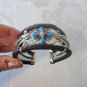 May include: A brown leather cuff bracelet with a silver and gold filigree design. The bracelet features a central cross-shaped design with turquoise-colored stones. The leather is braided, and the metal accents are detailed.