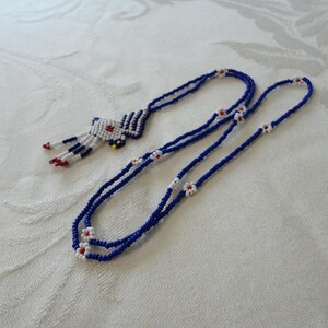 Pu&ograve; includere: Una collana di perline con un cordino blu e dettagli bianchi e rossi. La collana presenta un ciondolo con un design geometrico in bianco, blu, rosso e giallo. La collana &egrave; esposta su una superficie bianca.