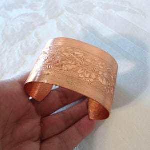 May include: Copper cuff bracelet with an embossed floral design. The wide band features detailed leaves and flowers. The cuff is held in a hand, showcasing the warm, metallic color and the intricate pattern. A vintage-inspired accessory.
