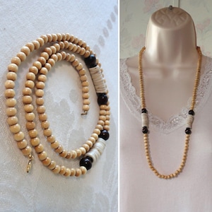 May include: A long, layered necklace featuring light brown wooden beads. The necklace includes dark brown and white accents. The necklace is displayed on a white surface and a mannequin.