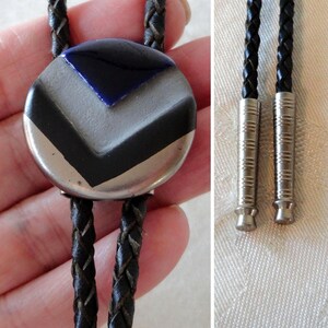 May include: A vintage bolo tie featuring a round silver-toned pendant with a geometric design in black, gray, and blue. The tie has a braided black leather cord with silver-toned metal tips. A classic accessory.