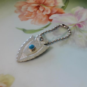 May include: A silver-toned metal arrowhead pendant attached to a small ball chain. The arrowhead features an engraved design of two crossed arrows and a central circular turquoise-colored stone set in a small metal bezel at the top center.
