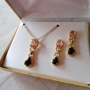 May include: A gold-toned jewelry set featuring a necklace and matching earrings, presented in a gift box. The necklace and earrings have a design with pink and black gemstones. The set is displayed on a white background.