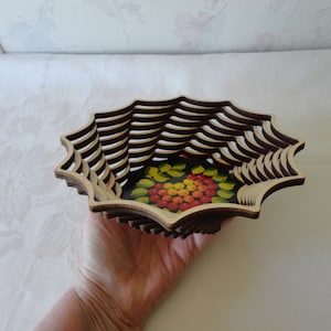 May include: A decorative wooden basket with a layered design. The basket has a dark brown rim and a light brown interior. The base features a painted floral design with red and yellow berries and green leaves.