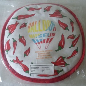 May include: A round white bread warmer with a red border, featuring red chili pepper illustrations. The center displays the text "BALLOON MUSEUM" in various colors, with additional text below. The warmer is 12 inches in diameter.