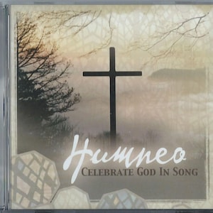 May include: A CD case featuring a serene landscape with a black cross as the focal point. The background has a soft, muted color palette with a misty effect. The text "Hymneo Celebrate God In Song" is written in a stylized font.