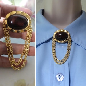 May include: A gold-tone brooch featuring an oval-shaped, dark brown cabochon stone set within a scalloped frame. Two delicate, textured gold-tone chains hang from the base of the brooch, creating a layered, dangling effect that adds movement.