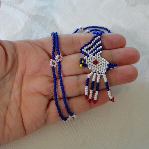 Pu&ograve; includere: Una collana e un ciondolo di perline con un design a forma di uccello. La collana &egrave; realizzata con piccole perline blu con accenti bianchi e rossi. Il ciondolo a forma di uccello &egrave; bianco con strisce blu, un centro rosso e dettagli rossi e gialli.
