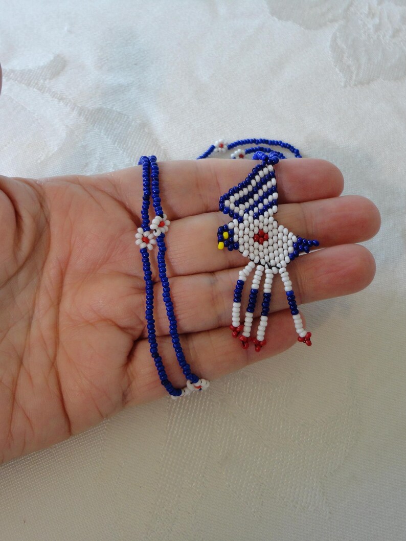 Pu&ograve; includere: Una collana e un ciondolo di perline con un design a forma di uccello. La collana &egrave; realizzata con piccole perline blu con accenti bianchi e rossi. Il ciondolo a forma di uccello &egrave; bianco con strisce blu, un centro rosso e dettagli rossi e gialli.