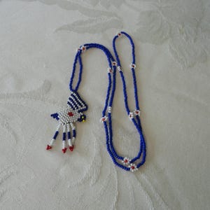 Pu&ograve; includere: Una collana di perline con una combinazione di colori blu, bianco e rosso. La collana presenta un ciondolo a forma di uccello e un doppio filo di perline con dettagli floreali bianchi e rossi. La collana &egrave; esposta su una superficie bianca.