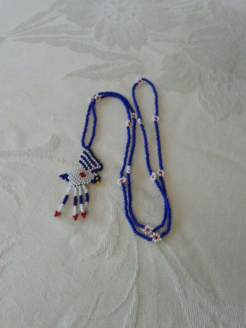 Pu&ograve; includere: Una collana di perline con una combinazione di colori blu, bianco e rosso. La collana presenta un ciondolo a forma di uccello e un doppio filo di perline con dettagli floreali bianchi e rossi. La collana &egrave; esposta su una superficie bianca.