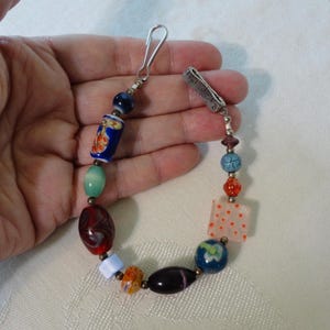 Puede incluir: Una colorida pulsera de cuentas con una variedad de cuentas de vidrio en tonos azules, rojos, naranjas y verdes. La pulsera incluye cuentas redondas, ovaladas y cuadradas, con un cierre plateado.