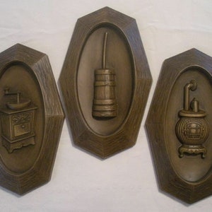 May include: Set of three brown wall plaques with relief images. One plaque features a coffee grinder, another a butter churn, and the third a wood-burning stove. Each image is set within an oval, framed by a textured, geometric border.