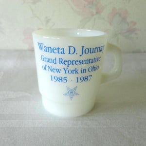 May include: A white ceramic mug with a handle. Blue text reads "Waneta D. Journay Grand Representative of New York in Ohio 1985-1987" with a star emblem. The mug has a slightly flared shape.