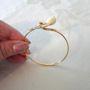 May include: A gold-colored bangle bracelet featuring a white flower bud design. The bracelet has a delicate, curved stem that wraps around the band. The flower bud is a creamy white color, adding a touch of elegance to the jewelry.