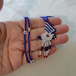 Pu&ograve; includere: Una collana di perline fatta a mano con un ciondolo a forma di uccello. Il ciondolo &egrave; bianco, blu e rosso, con un accento giallo. La catena della collana &egrave; blu con accenti floreali bianchi e rossi. La collana &egrave; tenuta in una mano.