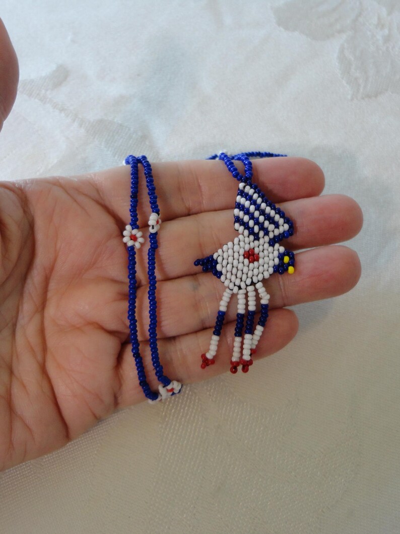 Pu&ograve; includere: Una collana di perline fatta a mano con un ciondolo a forma di uccello. Il ciondolo &egrave; bianco, blu e rosso, con un accento giallo. La catena della collana &egrave; blu con accenti floreali bianchi e rossi. La collana &egrave; tenuta in una mano.