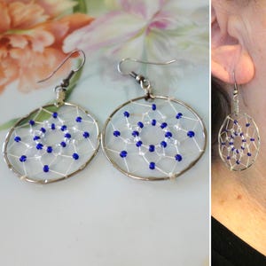 May include: A pair of circular drop earrings featuring a silver-toned metal hoop frame with a webbed design made of white thread. The web is accented with small, alternating royal blue and clear glass beads arranged in a geometric pattern.