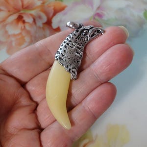 May include: A pendant featuring a cream-colored, curved tooth with a silver-colored eagle head. The eagle head has detailed feather and eye carvings. The pendant is suspended from a small silver loop.