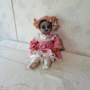 May include: A vintage doll with a pale face and dark eyes, wearing a pink dress with white lace trim and a matching bonnet. The doll is seated on a white surface, creating a spooky aesthetic.