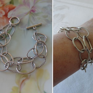 May include: A silver-toned bracelet featuring a series of interconnected oval and twisted links. The bracelet has a toggle clasp closure. The jewelry is displayed on a floral background and also on a wrist.
