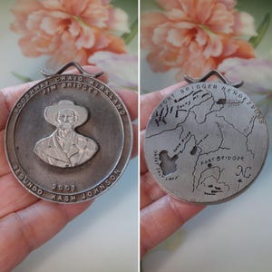 May include: A silver-colored medallion with a portrait of a man in a hat and the text "BOOSHWAY CRAIG KIRKLAND JIM BRIDGER SEGUNDO KASH JOHNSON 2001". The reverse side features a map with the text "FORT BRIDGER RENDEZVOUS".