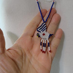 Pu&ograve; includere: Una collana di perline con un ciondolo a forma di uccello. Il ciondolo &egrave; bianco con dettagli blu e rossi. La collana ha una catena di perline blu con accenti floreali bianchi e rossi.