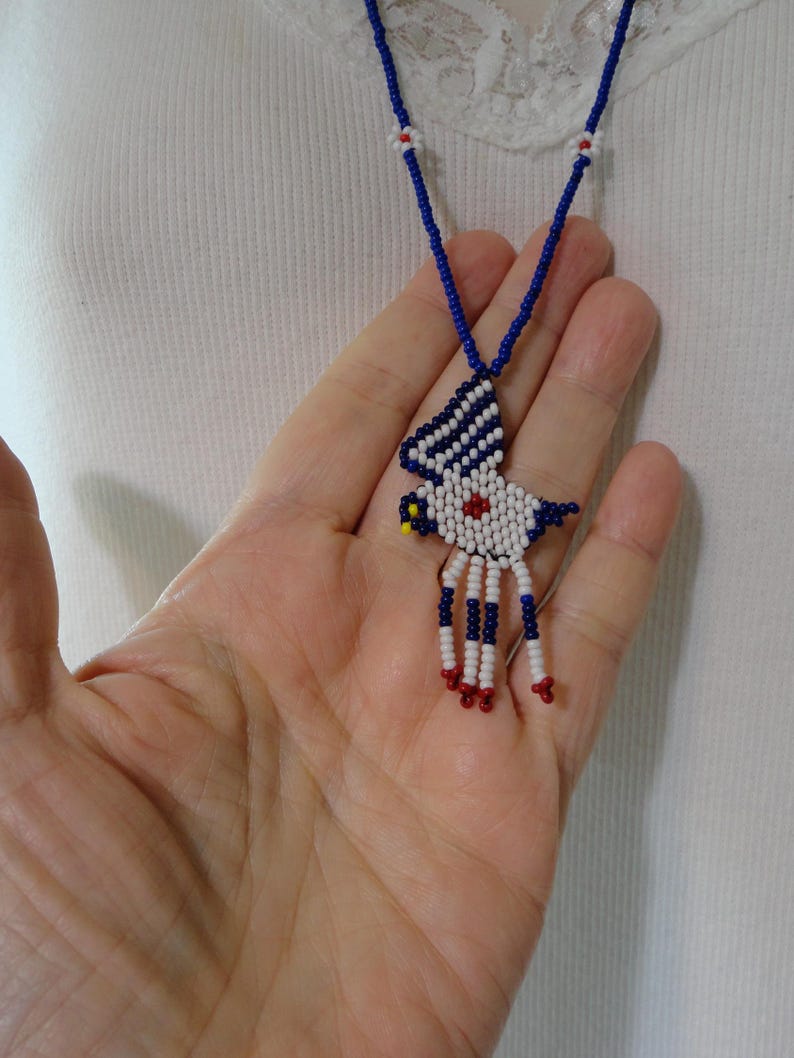 Pu&ograve; includere: Una collana di perline con un ciondolo a forma di uccello. Il ciondolo &egrave; bianco con dettagli blu e rossi. La collana ha una catena di perline blu con accenti floreali bianchi e rossi.