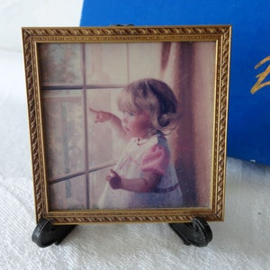 May include: A small, square picture frame with a gold-colored, ornate border. The frame holds a photograph of a young child in a pink dress, pointing towards a window. The frame is supported by a black easel.