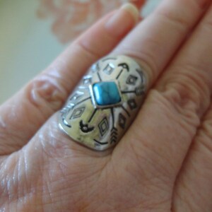 May include: Silver-toned ring with a turquoise-colored square stone at the center. The ring features an engraved design of arrows and geometric shapes. The ring is worn on a finger.