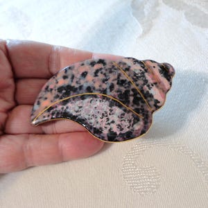May include: A seashell-shaped brooch with a speckled pattern in pink, black, and white. The brooch has a gold-colored trim and a glossy finish. The design is reminiscent of a natural seashell.