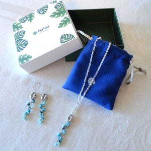 May include: A jewelry set featuring a silver necklace and matching earrings with turquoise-colored beads. The necklace and earrings are displayed with a blue velvet pouch and a white box with green leaf designs. The box has the word "Bahia" printed on it.