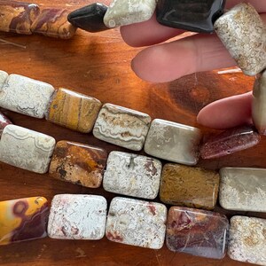 May include: A close-up of a necklace or bracelet featuring rectangular and rounded beads in various colors and patterns. The beads include shades of brown, beige, white, and black, with unique veining and textures. The jewelry is displayed on a wooden surface.