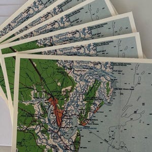 May include: A stack of greeting cards featuring a vintage map design. The maps have green, blue, and tan tones, with text indicating locations like Wolf Island National Wildlife Refuge and Altamaha River.