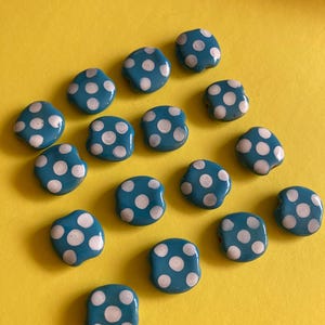 May include: A collection of turquoise blue, square-shaped beads with white polka dots. The beads are scattered on a bright yellow background, creating a playful and colorful display. These beads are ideal for jewelry making or craft projects.