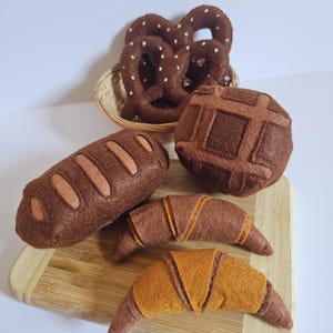 Könnte beinhalten: Eine Sammlung von Filz-Spielzeuglebensmitteln, bestehend aus zwei dunkelbraunen Brezeln in einem kleinen geflochtenen Korb, einem dunkelbraunen Brotlaib mit hellen Akzenten, einem runden dunkelbraunen Brötchen und zwei halbmondförmigen Croissants in Braun und Goldgelb.