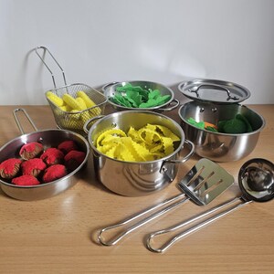 Könnte beinhalten: Ein Set aus Spielzeug-Kochgeschirr und -Utensilien aus Edelstahl. Das Set enthält Töpfe, Pfannen, einen Frittierkorb, einen Spatel und eine Kelle. Die Töpfe enthalten Spielzeug-Lebensmittel wie gelbe Nudeln, grünes Gemüse und rote Fleischbällchen.