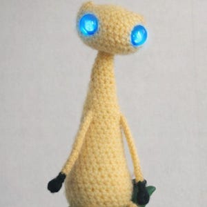 May include: A handmade, crocheted alien figure in light yellow yarn. The alien has a slender neck, long arms and legs, and glowing blue eyes. The hands and feet are black. The figure is sitting on a green surface.