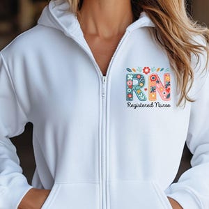 May include: A white zip-up hoodie featuring the letters "RN" in a floral design, with the words "Registered Nurse" below. The hoodie has a hood, a full-length zipper, and two front pockets. The design incorporates various colors, including blue, red, and yellow.