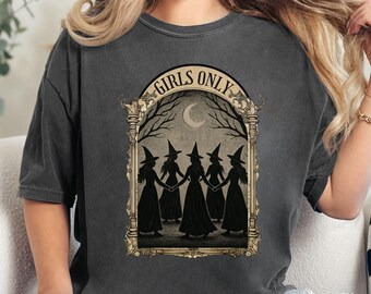 May include: Dark gray t-shirt featuring a vintage-style graphic of five witches holding hands under a crescent moon and bare tree branches. The text "GIRLS ONLY" is arched above the image in a gold frame.