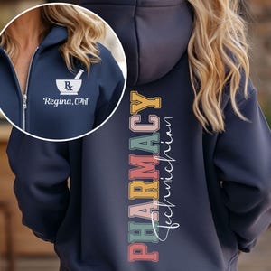 May include: Navy blue zip-up hoodie with "Pharmacy Technician" printed vertically on the back in colorful letters. A white mortar and pestle logo with the text "Regina, CPhT" is on the front. The hoodie has a hood and a silver zipper.