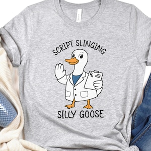 May include: A light grey t-shirt with a cartoon goose in a lab coat, holding a prescription pad. The text above the goose reads "SCRIPT SLINGING" and below it reads "SILLY GOOSE".