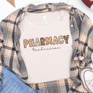 May include: A cream-coloured t-shirt with the words "PHARMACY technician" in a decorative, patterned font. The shirt is layered with a plaid button-down shirt in shades of grey, tan, and white. A football and trainers are also visible.