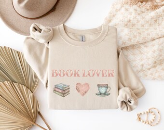 May include: A beige sweatshirt featuring the pink text "BOOK LOVER". The design includes illustrations of books, a heart, and a teacup. The sweatshirt is folded, with the sleeves visible.