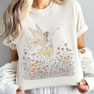 May include: Cream-colored t-shirt featuring a whimsical illustration of a fairy with outstretched arms, adorned with delicate wings and a yellow dress. The fairy is set amidst a field of colorful wildflowers, with the text "Women & Florals" at the bottom.