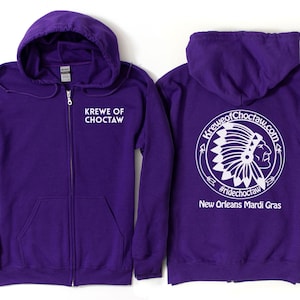 May include: A purple zip-up hoodie. The front displays the text "KREWE OF CHOCTAW". The back features a white circular design with a Native American chief illustration, the text "KREWEofChoctaw.com", "#ridechoctaw", and "New Orleans Mardi Gras".