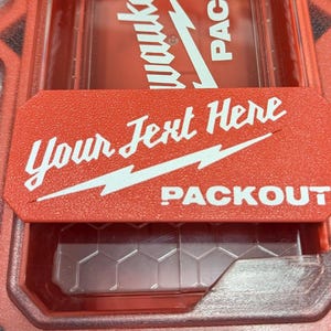 May include: Red PACKOUT storage container with a red lid. The lid has the text "Your Text Here" and "PACKOUT" in white. A white lightning bolt design is also on the lid.