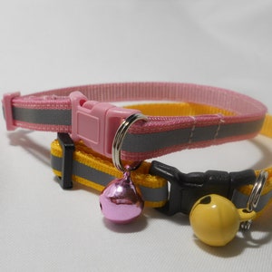 K&ouml;nnte beinhalten: Zwei Katzenhalsb&auml;nder, eines rosa und eines gelb, mit reflektierenden grauen Streifen. Jedes Halsband hat eine Kunststoffschnalle und eine kleine Glocke. Das rosa Halsband hat eine rosa Glocke, und das gelbe Halsband hat eine gelbe Glocke.