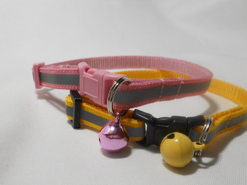 K&ouml;nnte beinhalten: Zwei Katzenhalsb&auml;nder, eines rosa und eines gelb, mit reflektierenden grauen Streifen. Jedes Halsband hat eine Kunststoffschnalle und eine kleine Glocke. Das rosa Halsband hat eine rosa Glocke, und das gelbe Halsband hat eine gelbe Glocke.