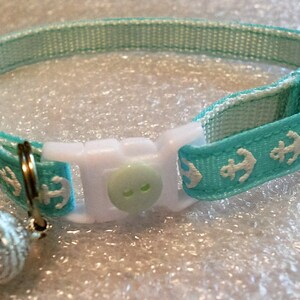 May include: A turquoise blue cat collar with white anchor designs and a white button. The collar has a silver bell attached.